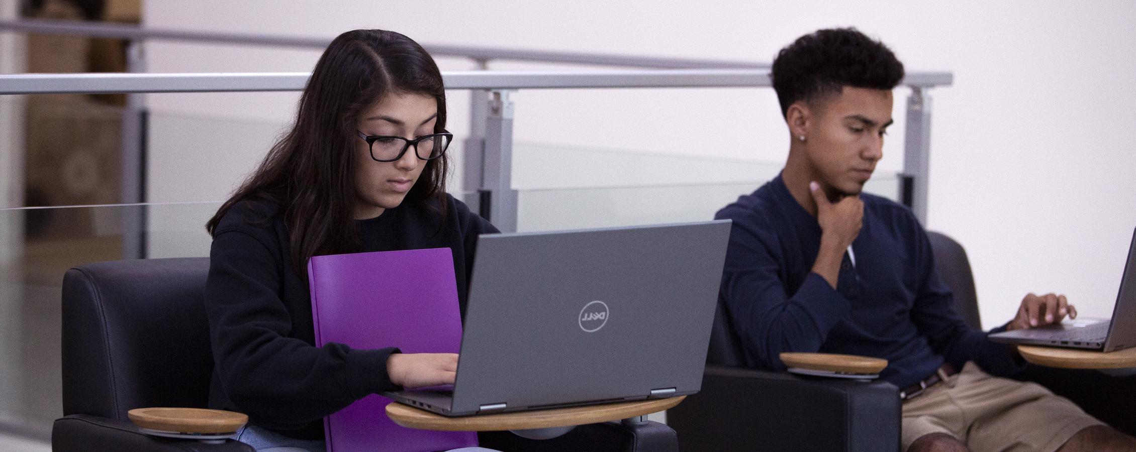 Dos estudiantes que trabajan con computadoras portátiles en un área de reunión.