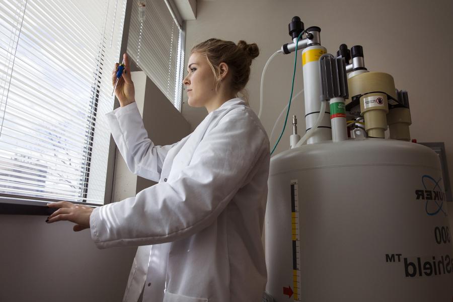 A student in a lab coat looks at a vile by the window.