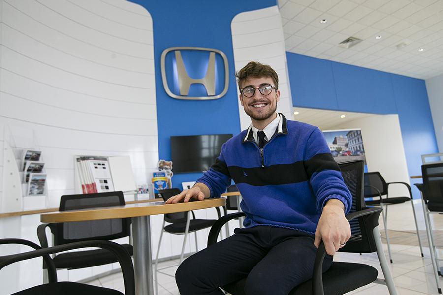 A student sits at a desk at a Honda dealership with a large Honda logo on the wall.