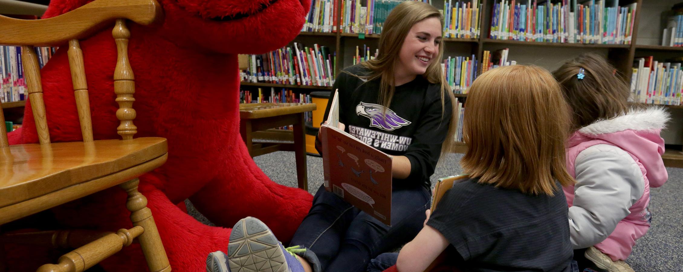 A Warhawk women's soccer player sits on the floor and reads with kids.