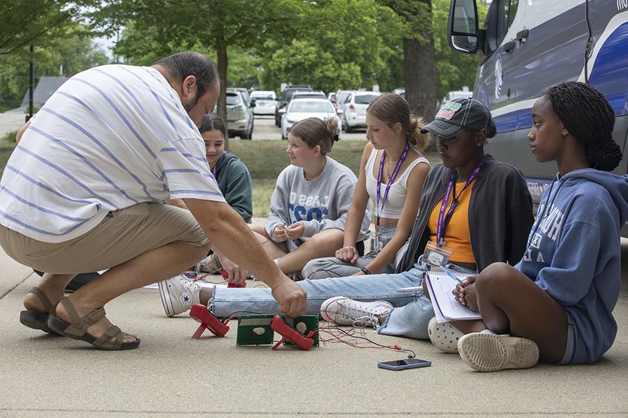 A faculty member kneels on the sidewalk with several precollege attendees, showing them electrical wiring.